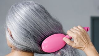 A woman with grey hair is picture brushing her hair with a pink hair brush