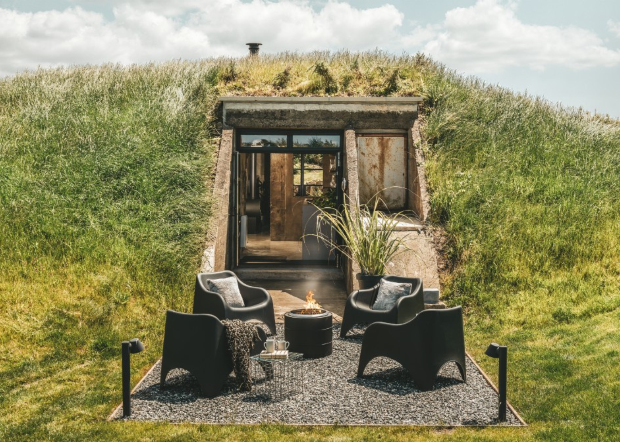 a world war 2 bunker has been redesigned as a luxury home, the building is concealed under a grassy hill, and there is a seating area with a firepit outside
