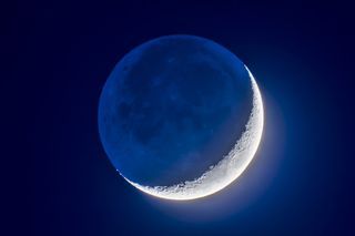 a thin crescent moon hangs in the night sky, while the "dark" portion of the moon's disk remains visible