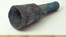 We see an old metal cannon near a ruler against a white background. One end of the cannon is wider than the other.