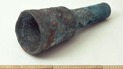 We see an old metal cannon near a ruler against a white background. One end of the cannon is wider than the other.
