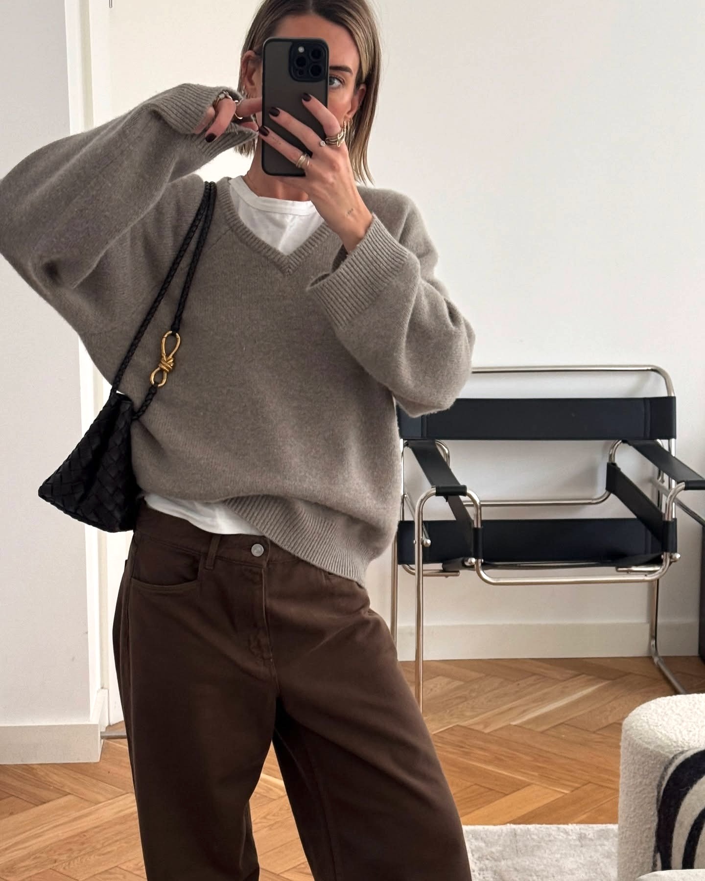 British influencer and stylist Hannah Lewis poses for a mirror selfie wearing a taupe V-neck sweater over a white T-shirt, a Bottega Veneta bag, and brown jeans