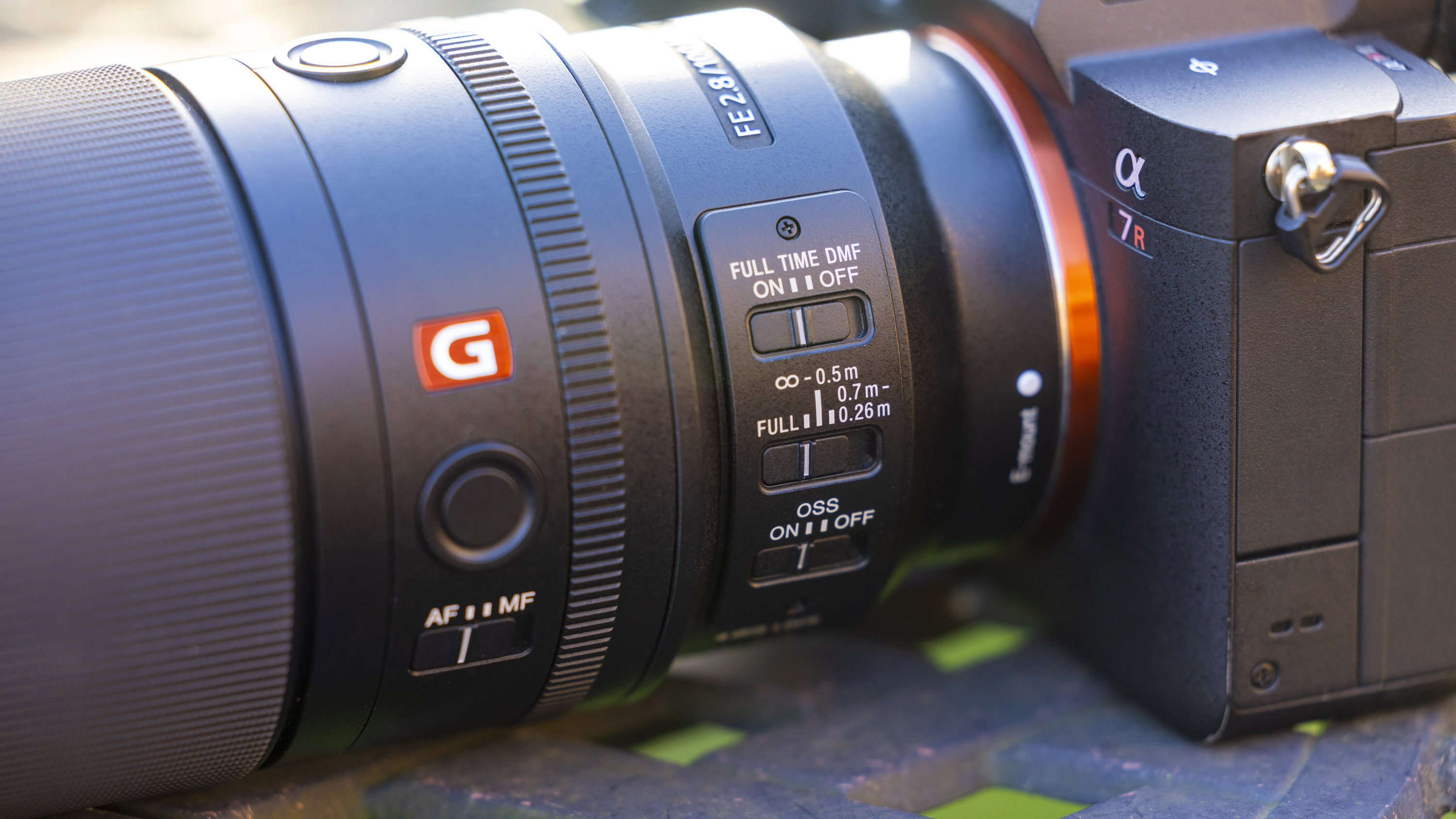 A product shot of the Sony FE 100mm f/2.8 Macro GM OSS attached to a Sony A7R IV outdoors on a metal garden table with a blurry background