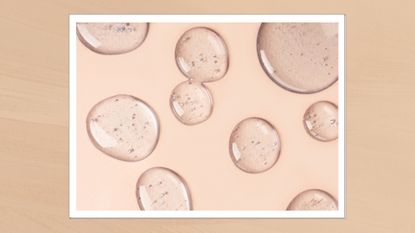 A close up of drops of a clear, serum-like liquid pictured on a pale pink backdrop/in a beige template