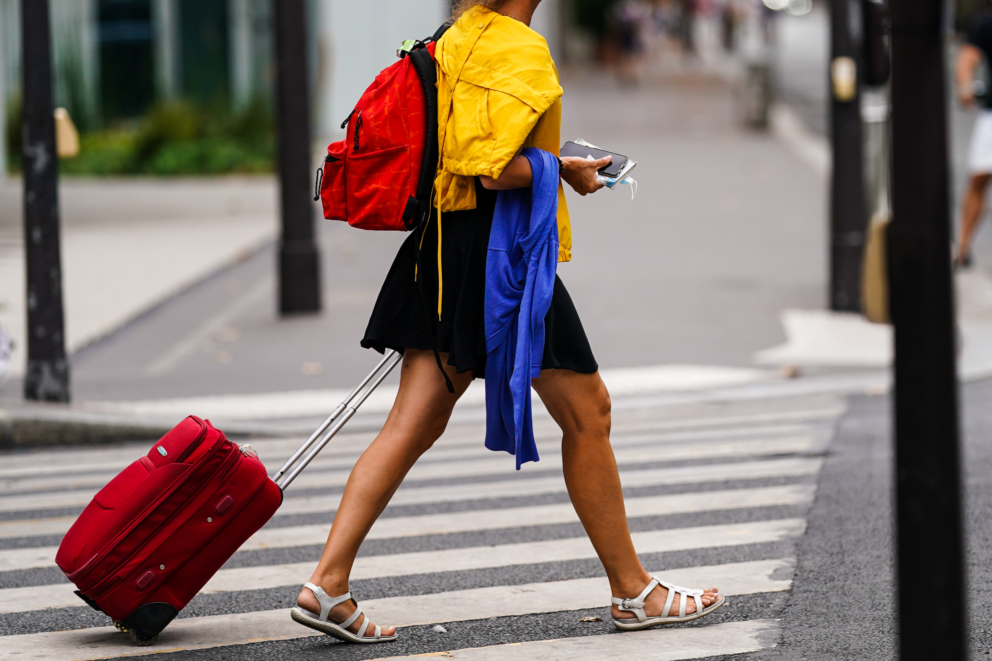 A person wearing a yellow jacket, black mini skirt, and white sandals, carrying a red backpack and rolling a red suitcase photographed on August 20, 2020 in Paris, France