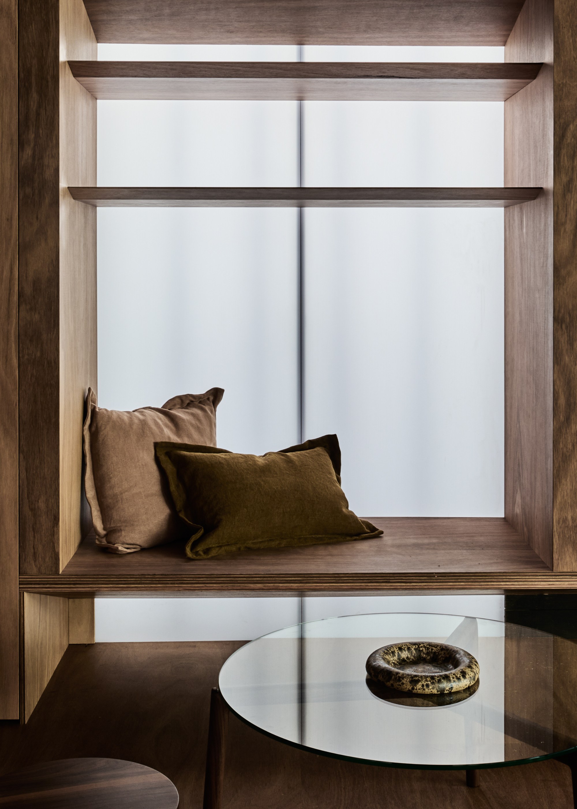 An entryway with in-built seating, a pair of pillows and a glass side table with a marble catchall