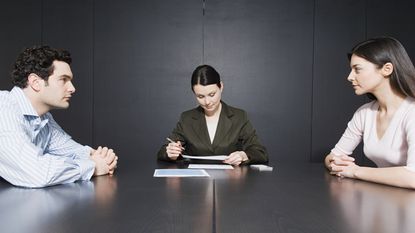 A divorcing couple sit on either side of a conference table with a lawyer between them looking at paperwork.