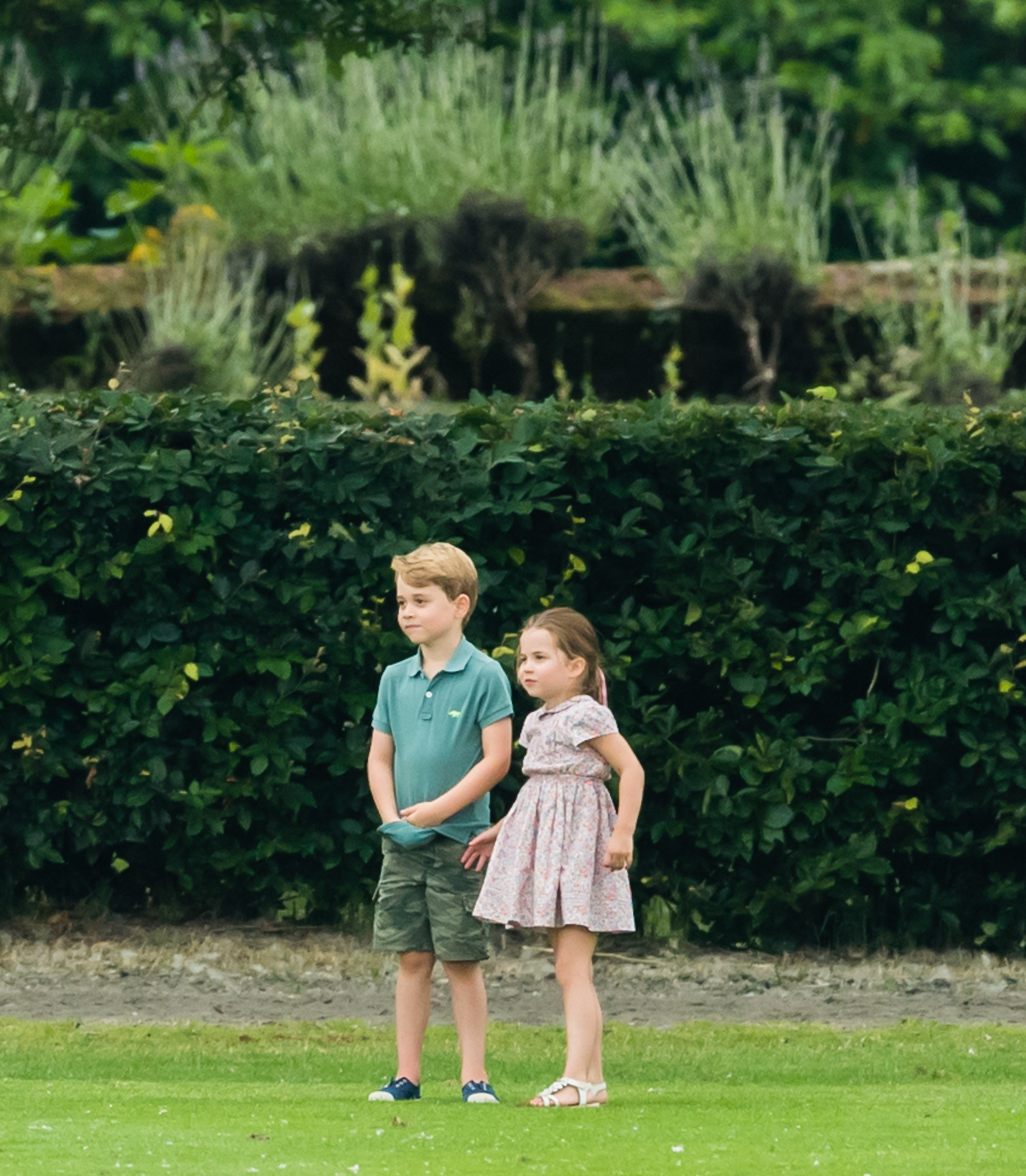 Prince George wearing shorts and a polo, and Princess Charlotte wearing a floral dress standing on a field