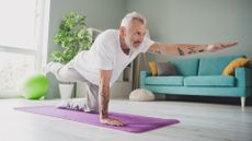 man wearing a white tshirt performing a bird dog exercise on a purple exercise mat. there's a turquoise sofa and green exercise ball behind him