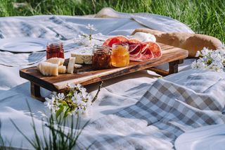 A grey chequered picnic blanket on the grass surrounded by bunches of white flowers, along with a wooden board with cheese, honey, meets, and jams laid out on top.