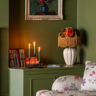 A colour-drenched green-painted living room with a fringed table lamp and lit candle sticks on a side cabinet