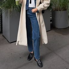 style influencer Coco Schiffer poses on a sidewalk in NYC while holding a green juice wearing a trench coat, straight-leg jeans and black ruched loafers