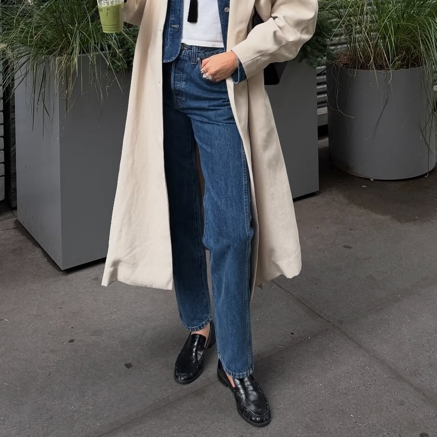 style influencer Coco Schiffer poses on a sidewalk in NYC while holding a green juice wearing a trench coat, straight-leg jeans and black ruched loafers