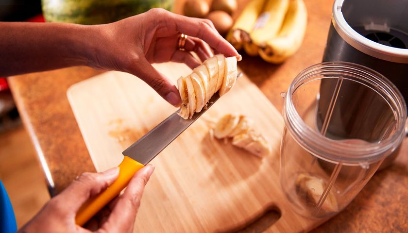 A woman&#039;s hands holding a knife with slices of banana on the blade by a smoothie jug