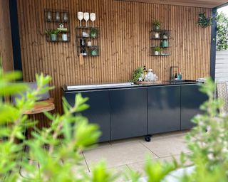Textured outdoor kitchen set-up with wood paneled wall, and black units on wheels.