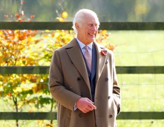 King Charles wearing a tan coat standing in front of a fence and a tree with gold and orange autumn leaves and laughing