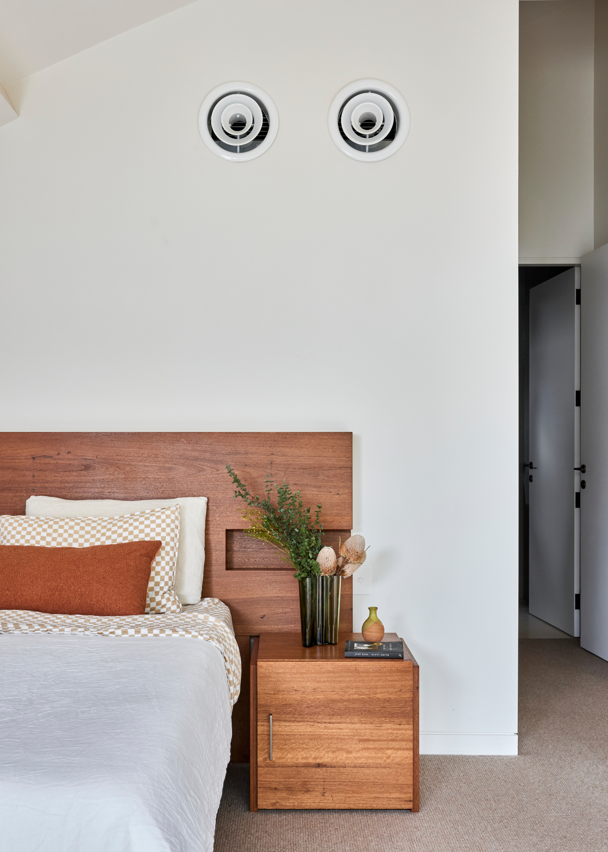 A white bedroom with a warm wood headboard and side table, a white bedscape with copper and checkerboard pillows