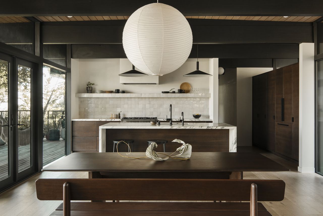 An open plan kitchen diner with a marble island, a large lamp shade, and a dark wooden dining bench 