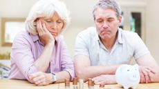 A worried senior couple sitting at a table looking at a piggy bank and several stacks of coins