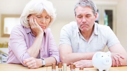 A worried senior couple sitting at a table looking at a piggy bank and several stacks of coins