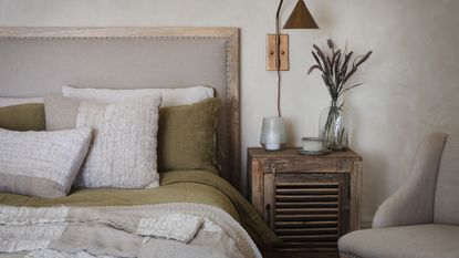 A bed with green linen bedding and a white throw pillow. A rustic wooden bedside table beside it with dried flowers in a vase