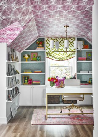 A maximalist home office with pink patterned wallpaper on the ceiling and open shelving