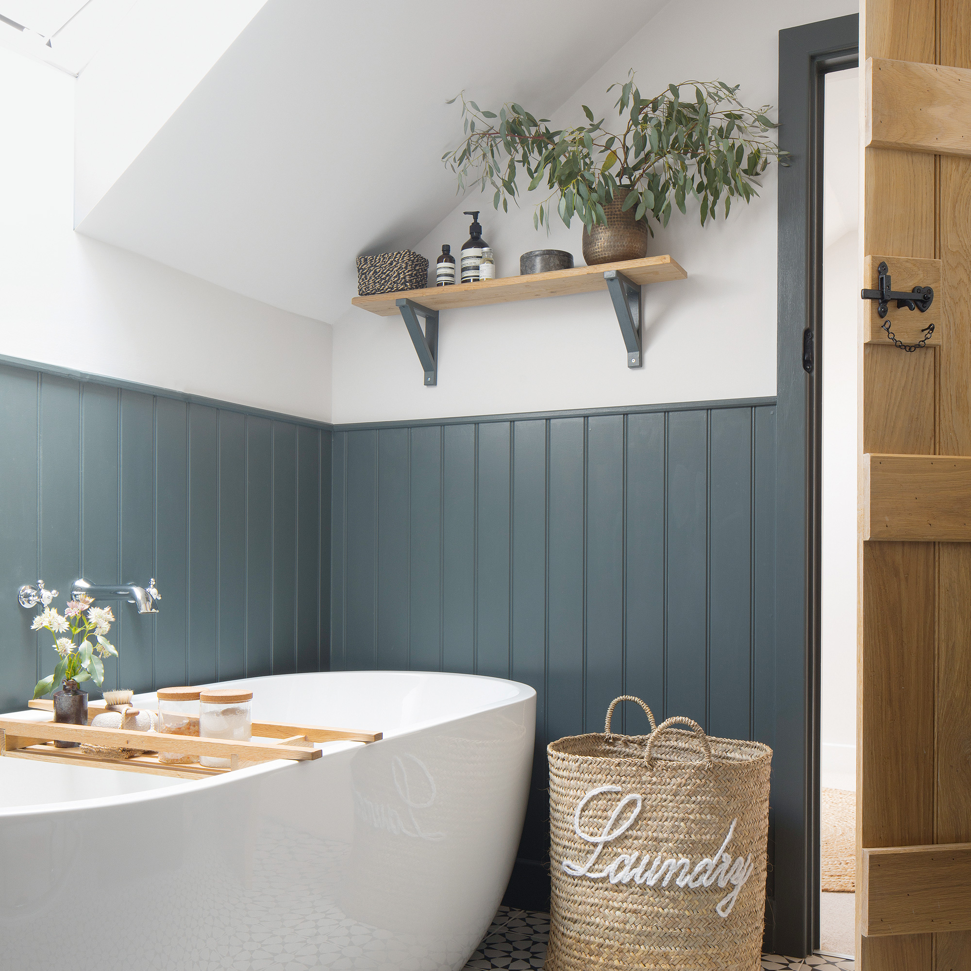 country cottage bathroom with tongue and grove panelling, a large contemporary bath, beside a wicker laundry basket and wall-hung shelving