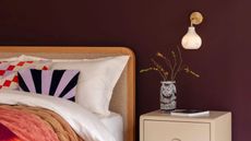 heal's wall sconce in bedroom with burgundy walls, fleece headboard, white sheets, colorful cushions, cream bedside table with colorful vase and book