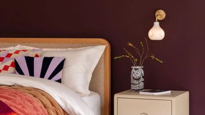 heal's wall sconce in bedroom with burgundy walls, fleece headboard, white sheets, colorful cushions, cream bedside table with colorful vase and book