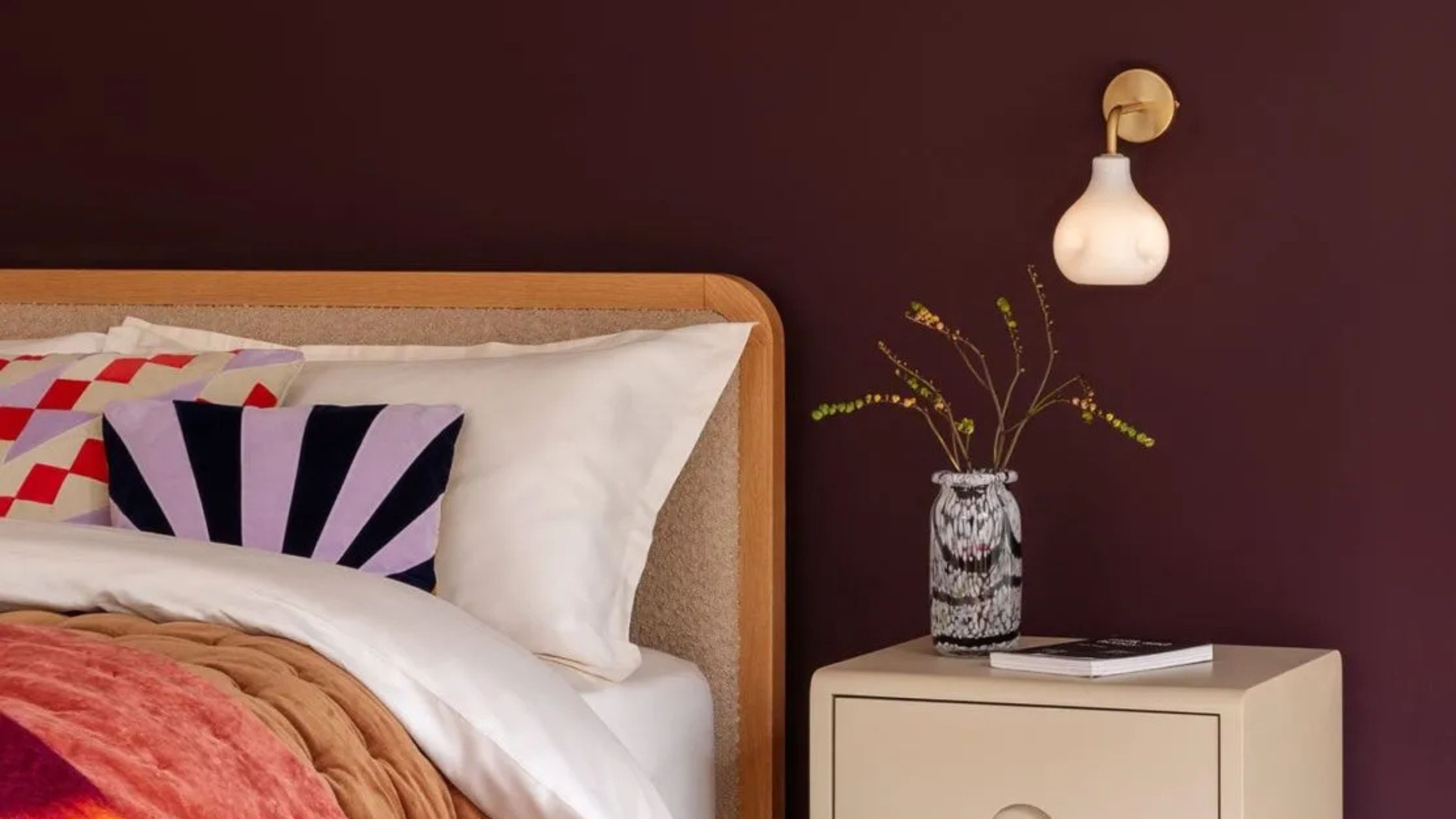 heal's wall sconce in bedroom with burgundy walls, fleece headboard, white sheets, colorful cushions, cream bedside table with colorful vase and book