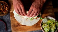 Shot from above of hands rolling a burrito on a wooden board. There is a bowl of chopped lettuce bottom right of image and a bowl of cooked meat in top left of image.