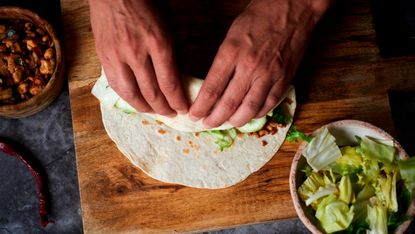 Shot from above of hands rolling a burrito on a wooden board. There is a bowl of chopped lettuce bottom right of image and a bowl of cooked meat in top left of image.