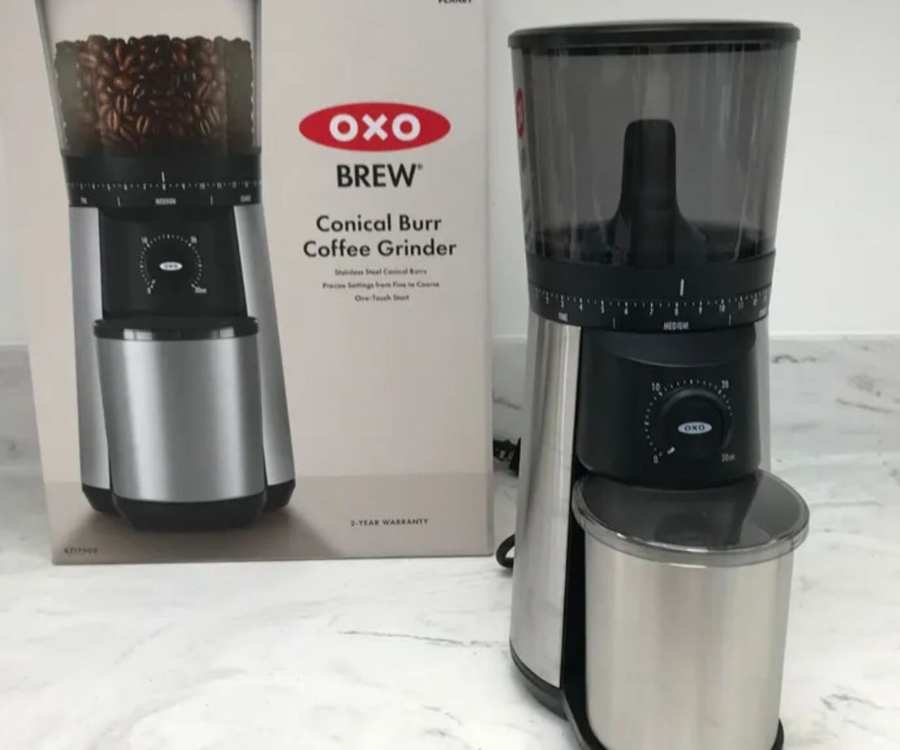 A coffee grinder with a stainless steel base and catch cup, with a black clear plastic hopper on top, in front of its branded OXO box, on a white marble counter top.