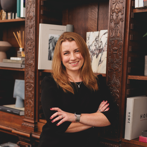 a headshot of ailsa connery, a woman with medium length light brown hair smiling at the camera with her arms crossed wearing a black dress and stading in front of an ornate dark wood display cabinet