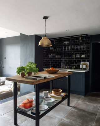 A kitchen with wall of black metro tiles, grey stone tile flooring and brass pendant ceiling light
