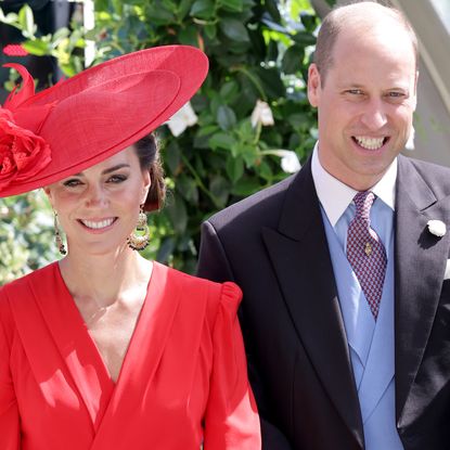 Kate Middleton wears a red hat with a wide brim and a matching red wrap dress, while Prince William wears a suit and waistcoat