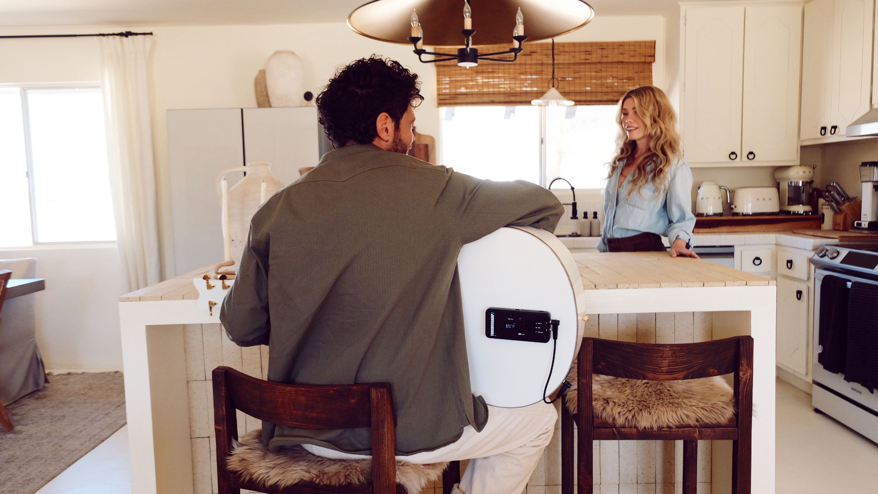 An image showing the ToneWoodAmp2 placed on the back of a white acoustic guitar, held by a man. He is sitting at a tall table in a white kitchen, and we see him from behind, allowing us to see the ToneWoodAmp2 in position. A woman stands opposite the table from the man, smiling at him, as if they are in conversation. &amp;quot;Hey, champ! That guitar sounds pretty good,&amp;quot; she seems to say. &amp;quot;Must be a ToneWoodAmp2 you&#039;ve got on there.&amp;quot;