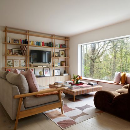 A living room with a large window, a grey sofa with a wooden frame, a brown settee-style sofa and an ottoman coffee table