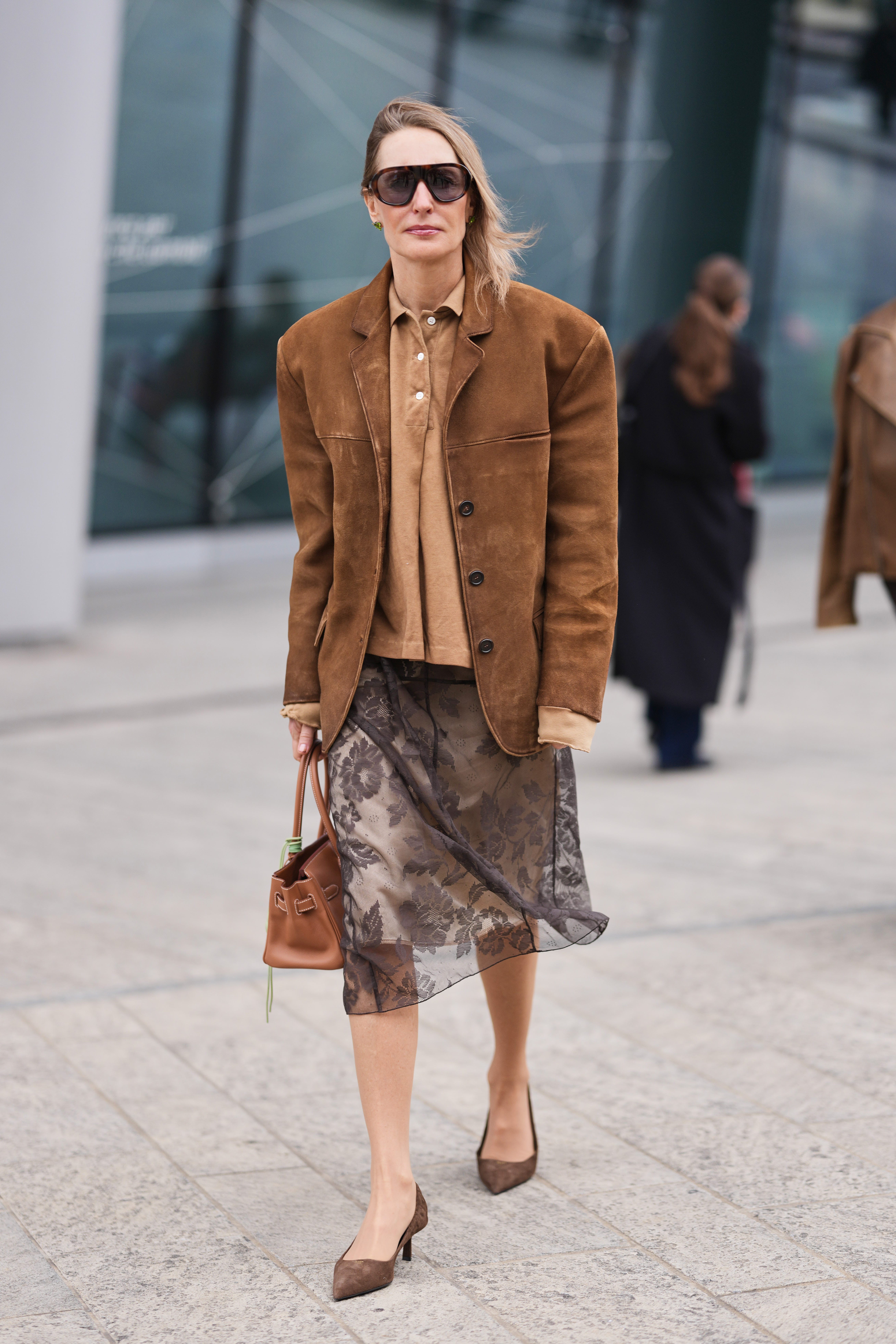 MILAN, ITALY - FEBRUARY 28: A guest wears dark brown large futurist sunglasses, a brown suede tailored jacket, a beige cotton button front shirt, a a beige cotton and black sheer floral embossed pattern knees skirt, a brown long leather Birkin handbag from Hermes with green pendant leather charms, brown suede pumps shoes from Prada, outside Ermanno Scervino, during Milan Fashion Week - Womenswear Fall/Winter 2026/2027, on February 28, 2026 in Milan, Italy (Photo by Edward Berthelot/Getty Images)