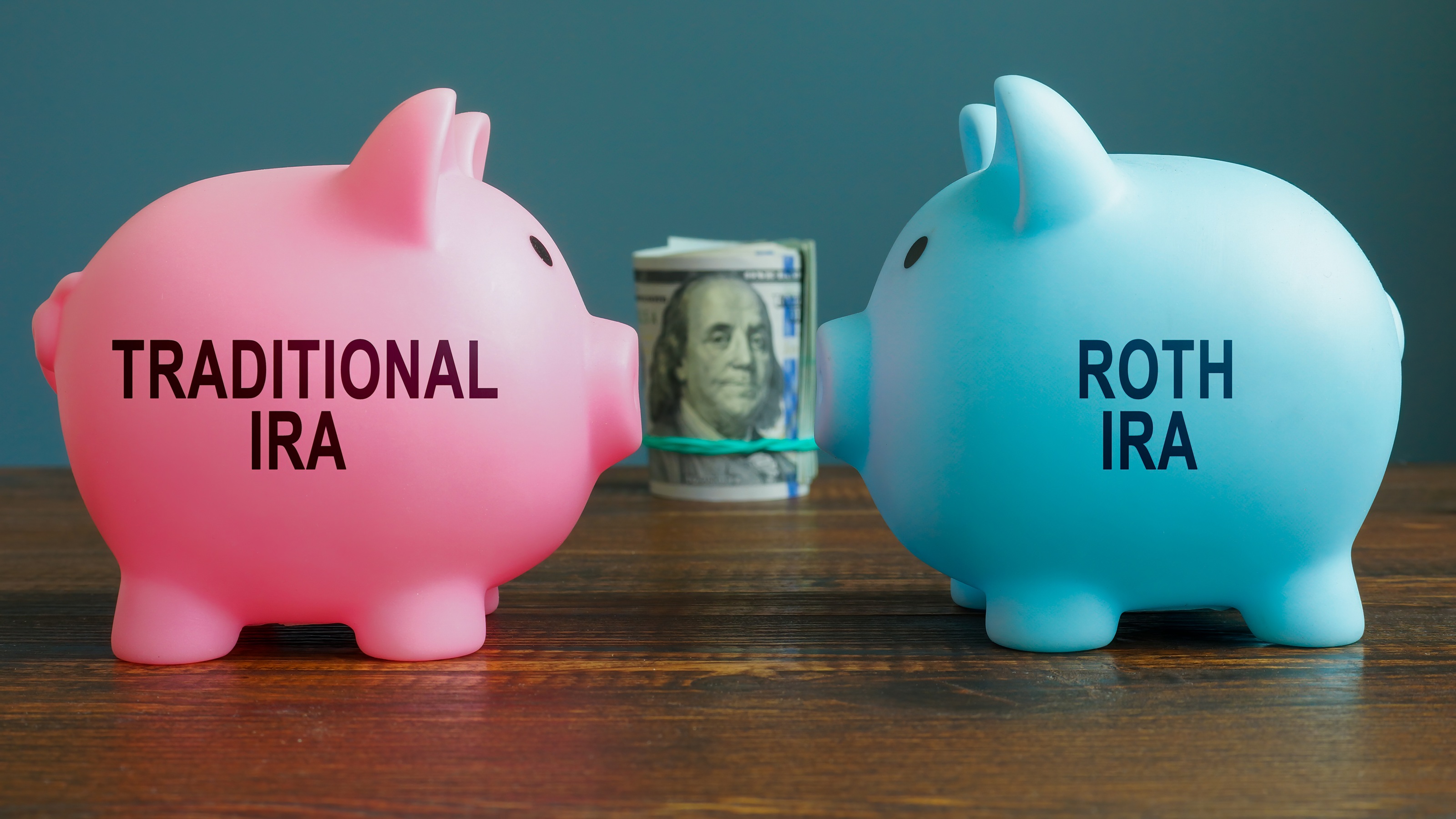 A pink piggy bank with "traditional IRA" on its side faces a blue piggy bank with "Roth IRA," a roll of hundreds between them.