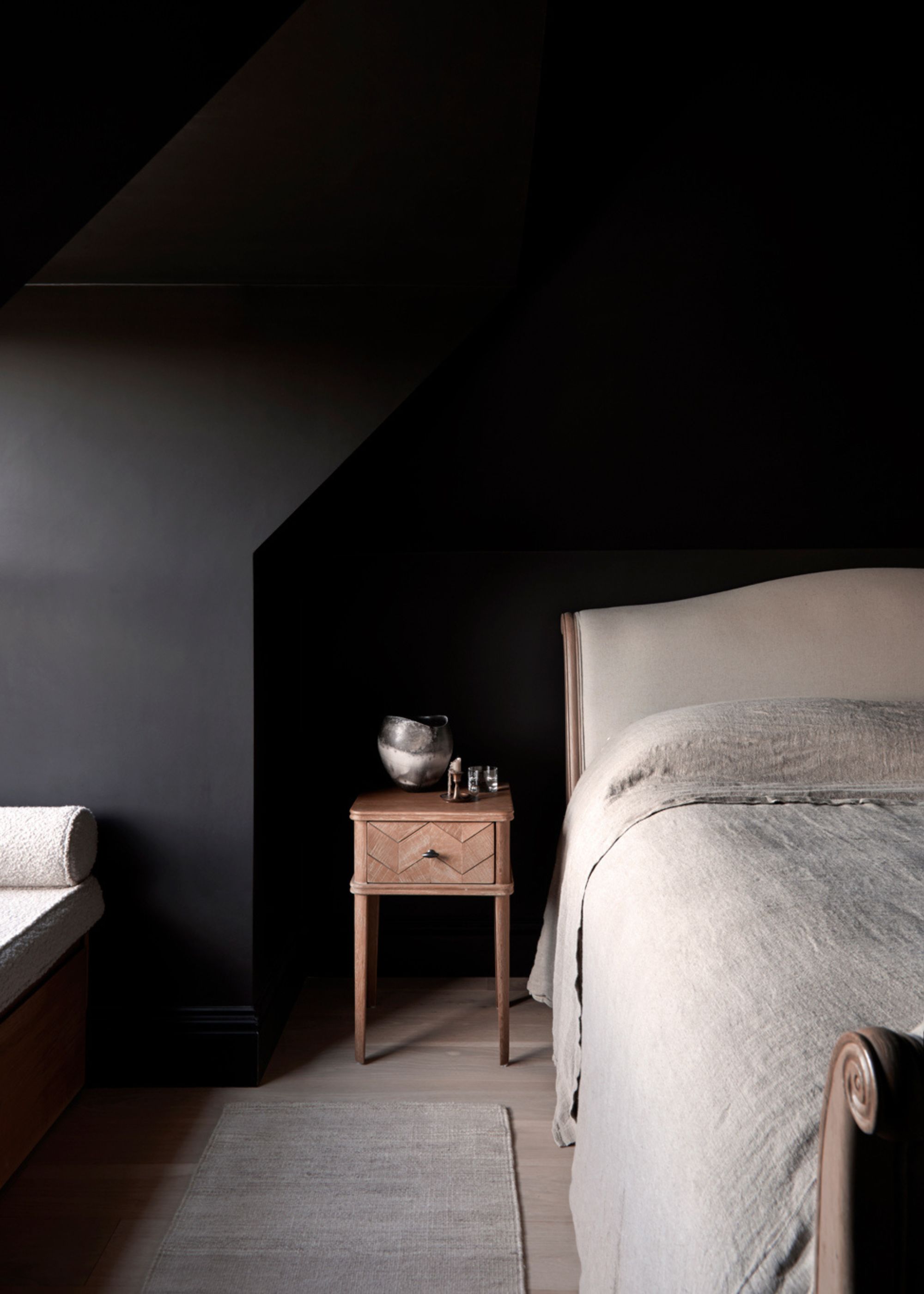 A dark bedroom with a beige bed, a wood nightstand, and a boucle cushioned bay window