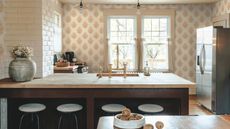 A small but bright kitchen with two windows, and peach wallpaper. There is a a wooden countertop on the kitchen peninsula, and a stainless steel fridge freezer in view. The ktichen has brass hardware and bar stools