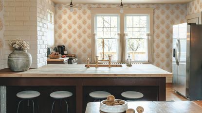 A small but bright kitchen with two windows, and peach wallpaper. There is a a wooden countertop on the kitchen peninsula, and a stainless steel fridge freezer in view. The ktichen has brass hardware and bar stools 