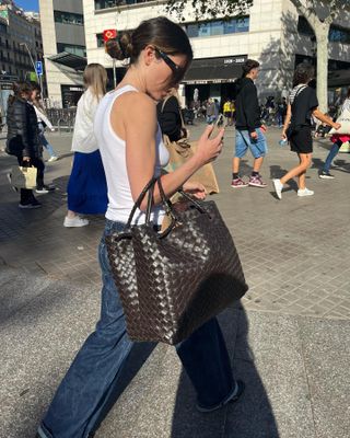 A woman wearing a white tank top, wide-leg jeans, sunglasses, and a large Bottega Veneta Andiamo bag.