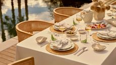 An outdoor table with a white cloth, rattan chargers, plates and wine glasses on top of it, with woven chairs next to it and a pool to the left of it