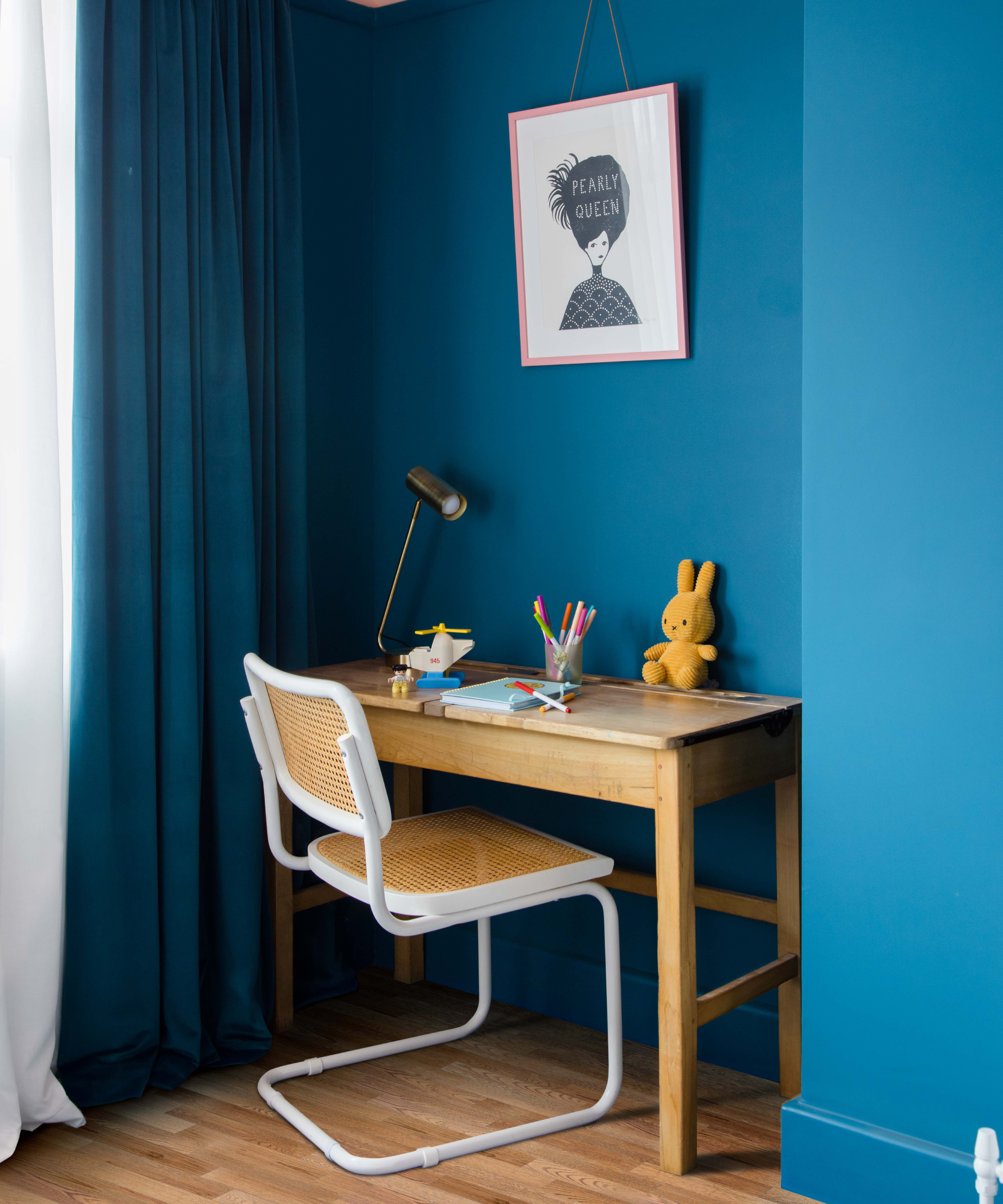 desk in alcove with walls painted blue, matching colour curtains, wooden desk, wicker and white desk chair and black metal task lamp on desk