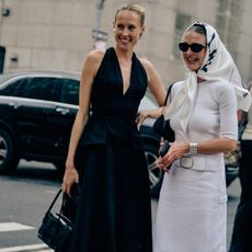 Two women at Fashion Week wearing a black dress and black bag and white top and skirt with a silk scarf