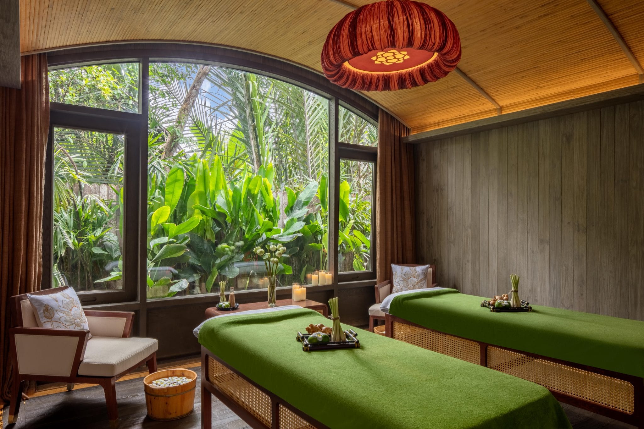A sage green and wood-tinted spa treatment room decorated with traditional Vietnamese furniture and filled with views of lush greenery.
