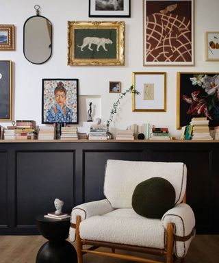 Gallery wall filled with art in different sized frames and mirrors, hung above black side board in front of a white boucle arm chair with a black cushion
