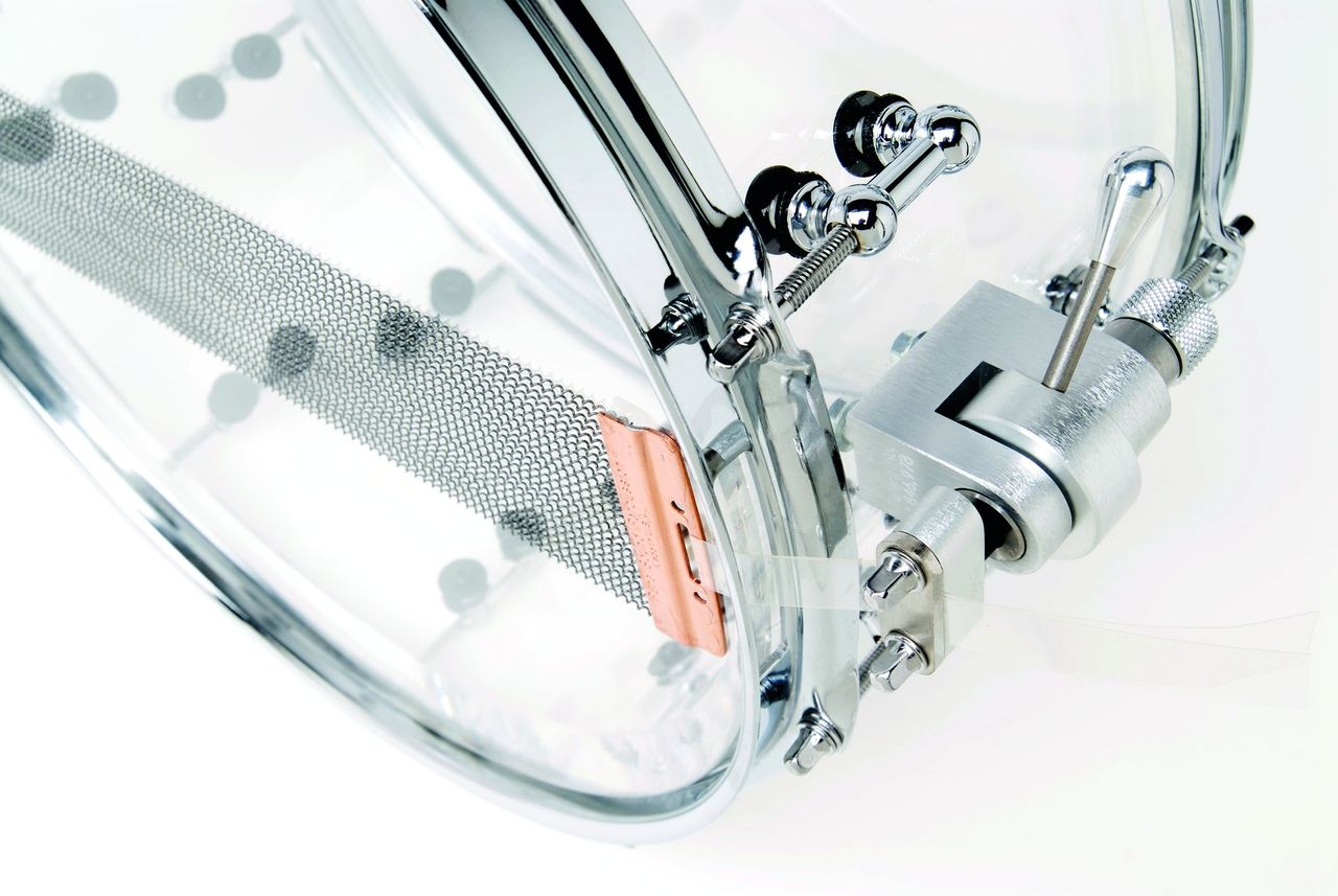 The clear shell snare drum has offset mini tube lugs and is fitted with Trick strainer and 24-strand Puresound snares